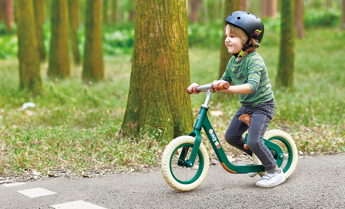A young child rides confidently through a forest path on the Balance Bike Bundle Green, one of the best children's outdoor toys for first-time riders.