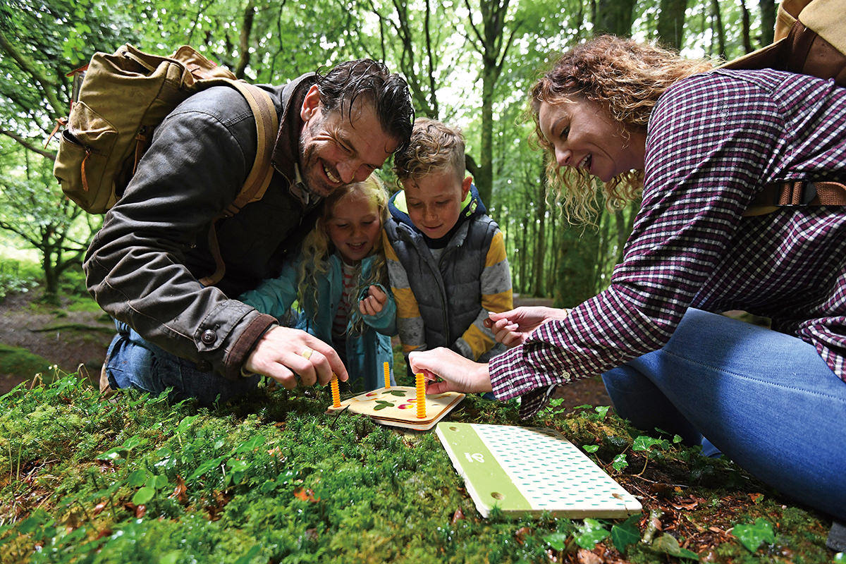 Family gathered in a forest playing a game on a mossy tree stump – a great example of toys for outside play that encourage teamwork and learning through.
